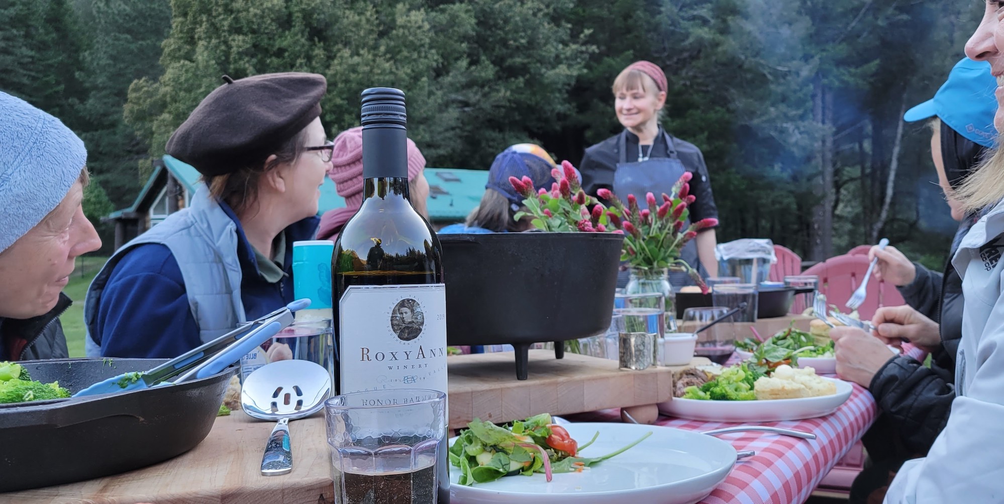 People around a wooden dining table outside eating dinner along the Rogue River with the chef at the head of the table standing up