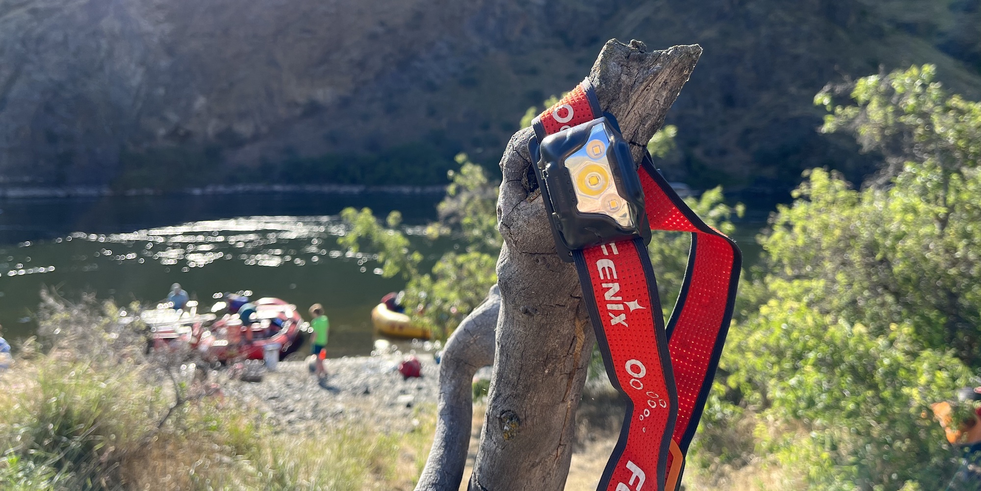A red and black headlamp hanging from a tree branch with the Snake River and red rafts blurred in the background