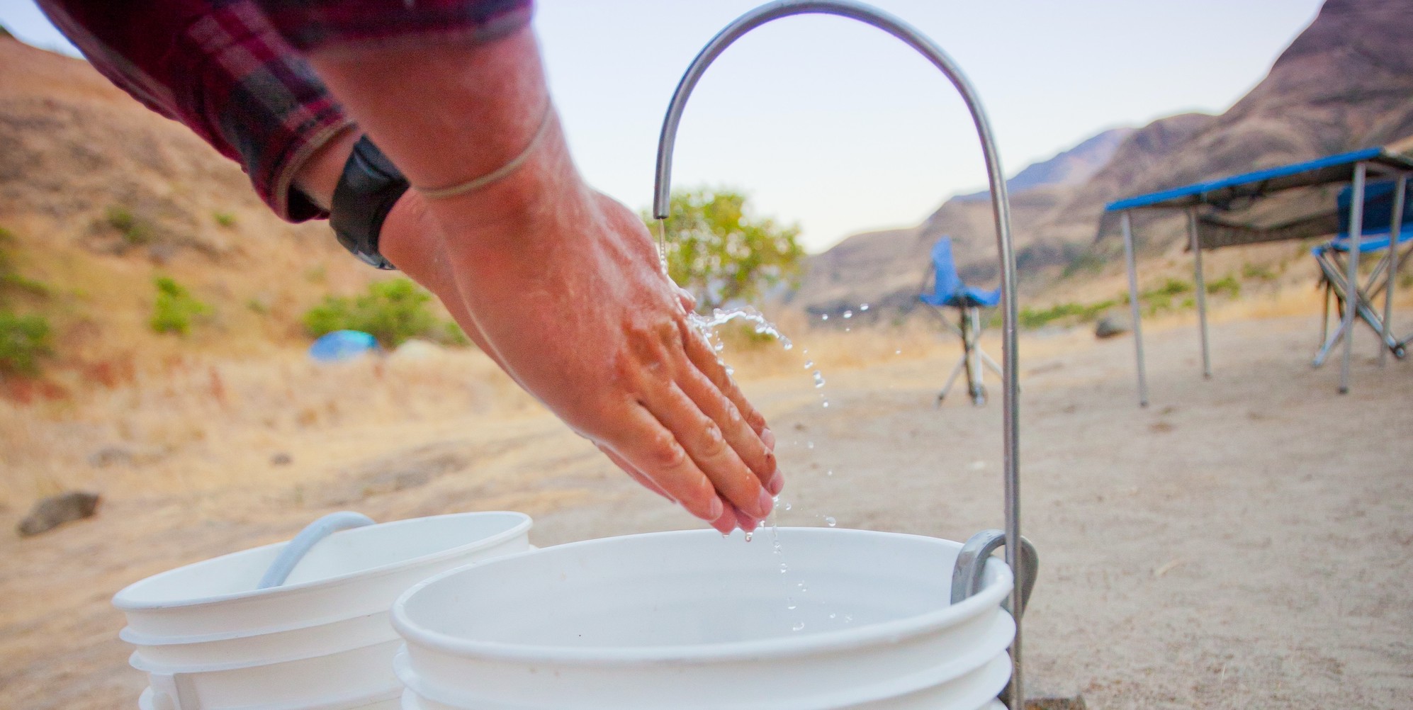 A person washing their hands into a white bucket while camping along a river