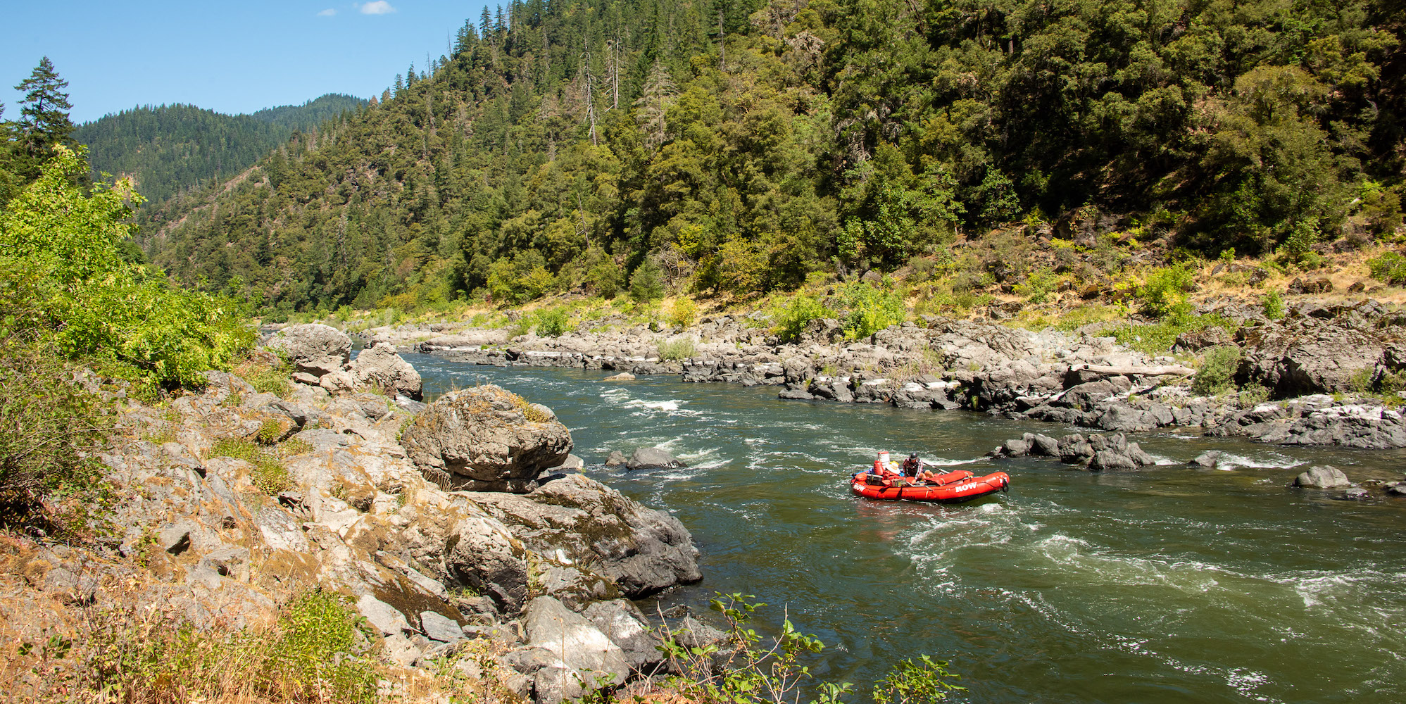 A red raft with one person in it floating downstream on the Rogue River