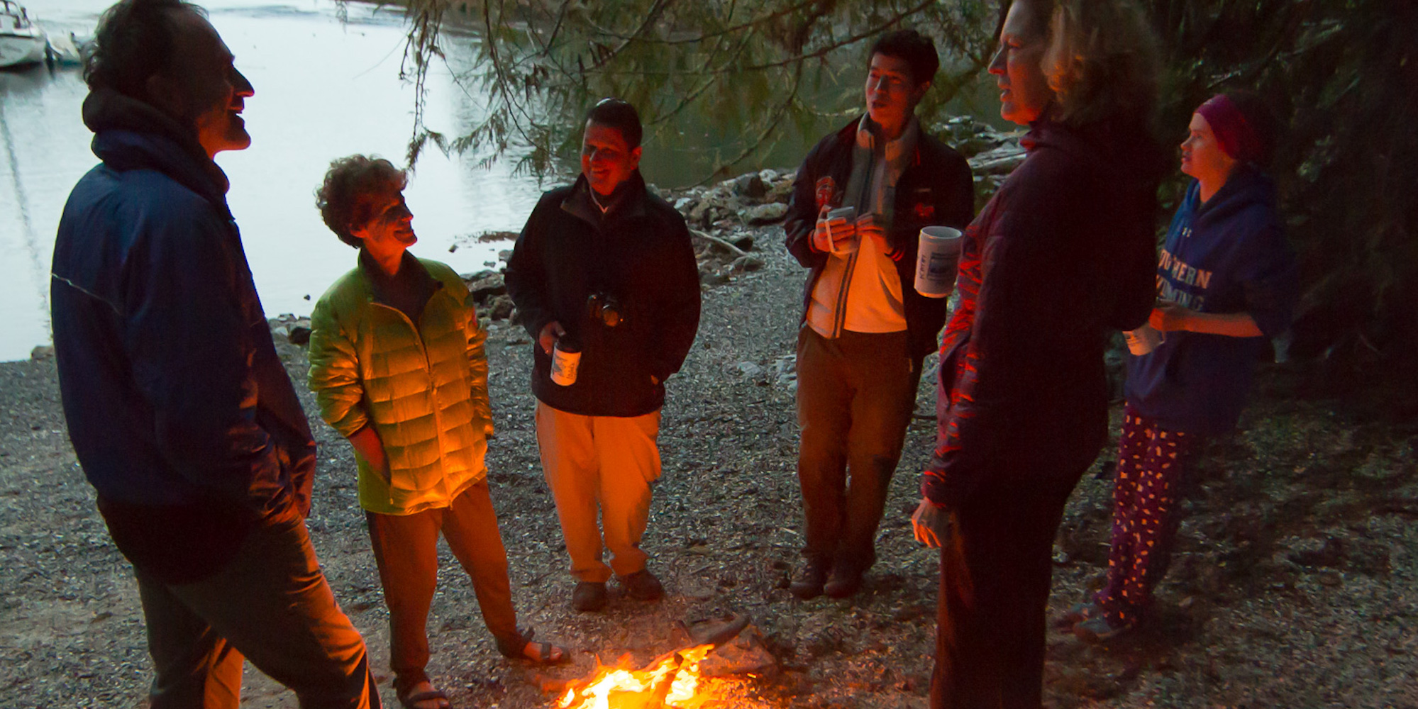 People in puffy jackets standing around a campfire on a rocky beach at sunset in British Columbia