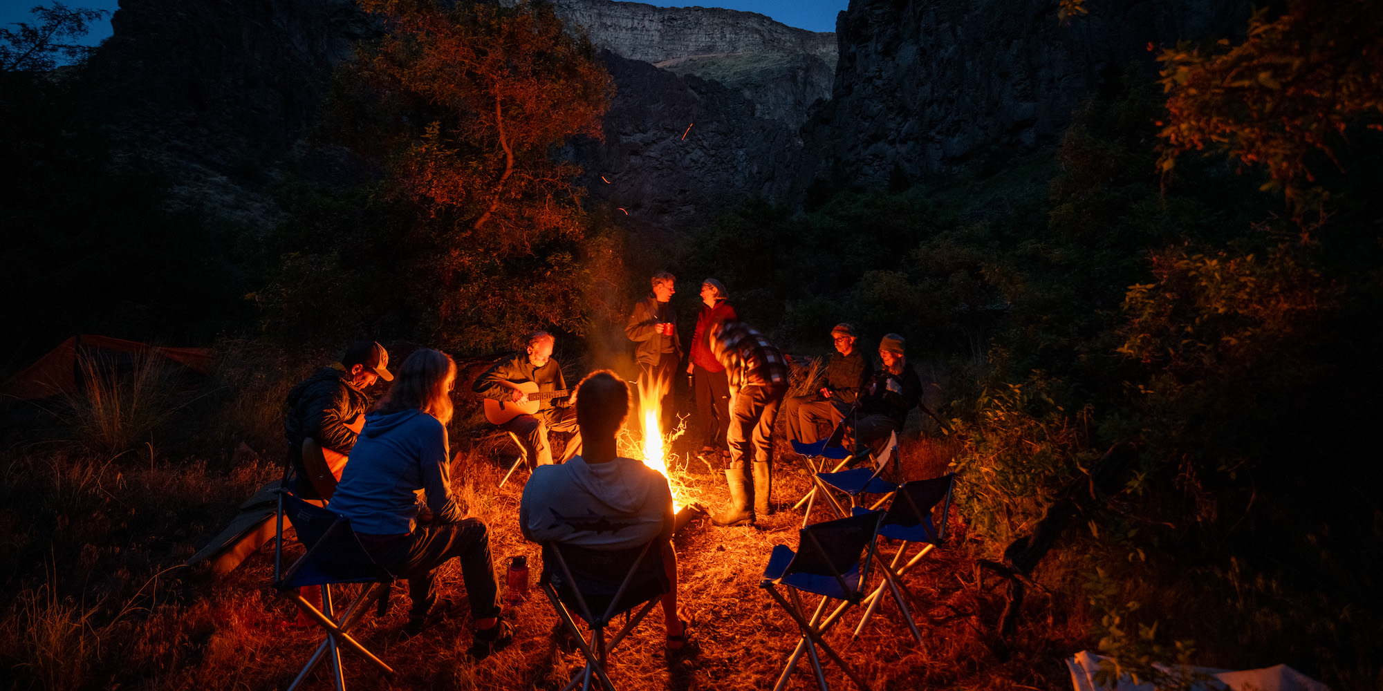 A group of people sitting around a campfire in Idaho talking amongst one another while one person plays guitar.