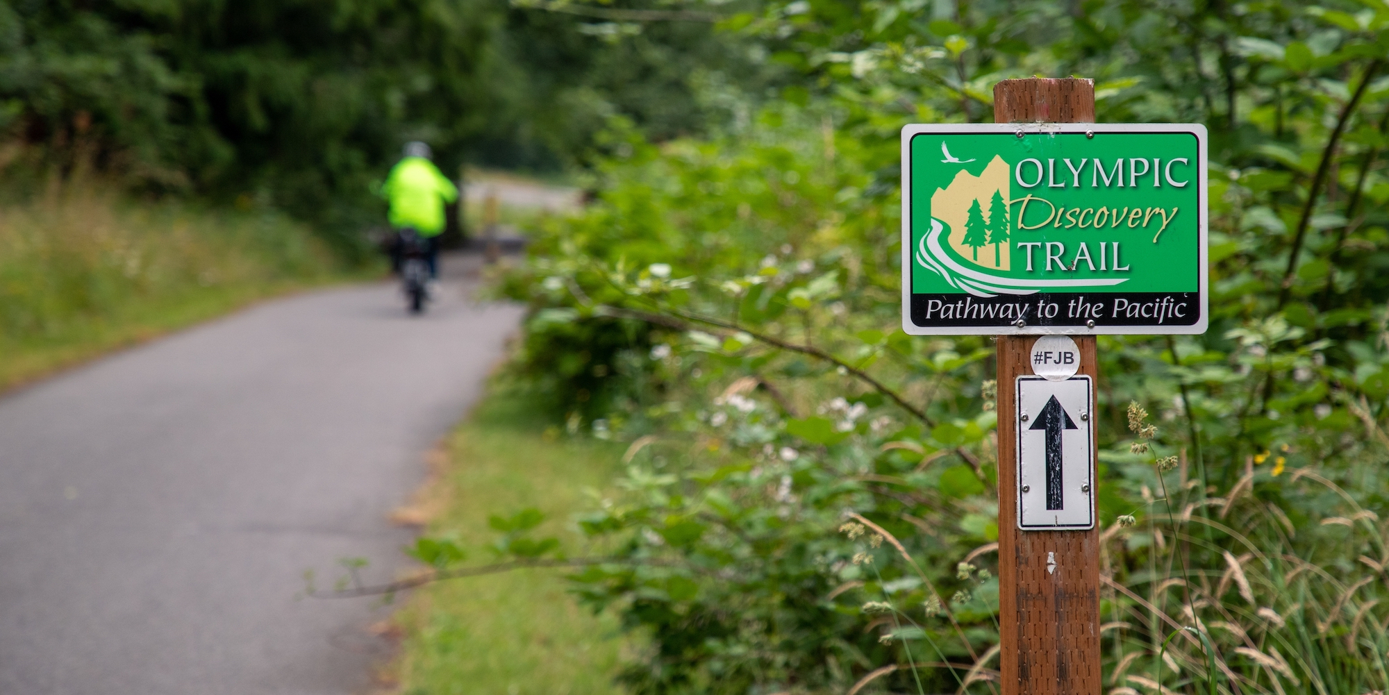 Sign post for the Olympic Discovery Trail using the rule of thirds with a person biking away on the trial in the blurred two thirds of the image
