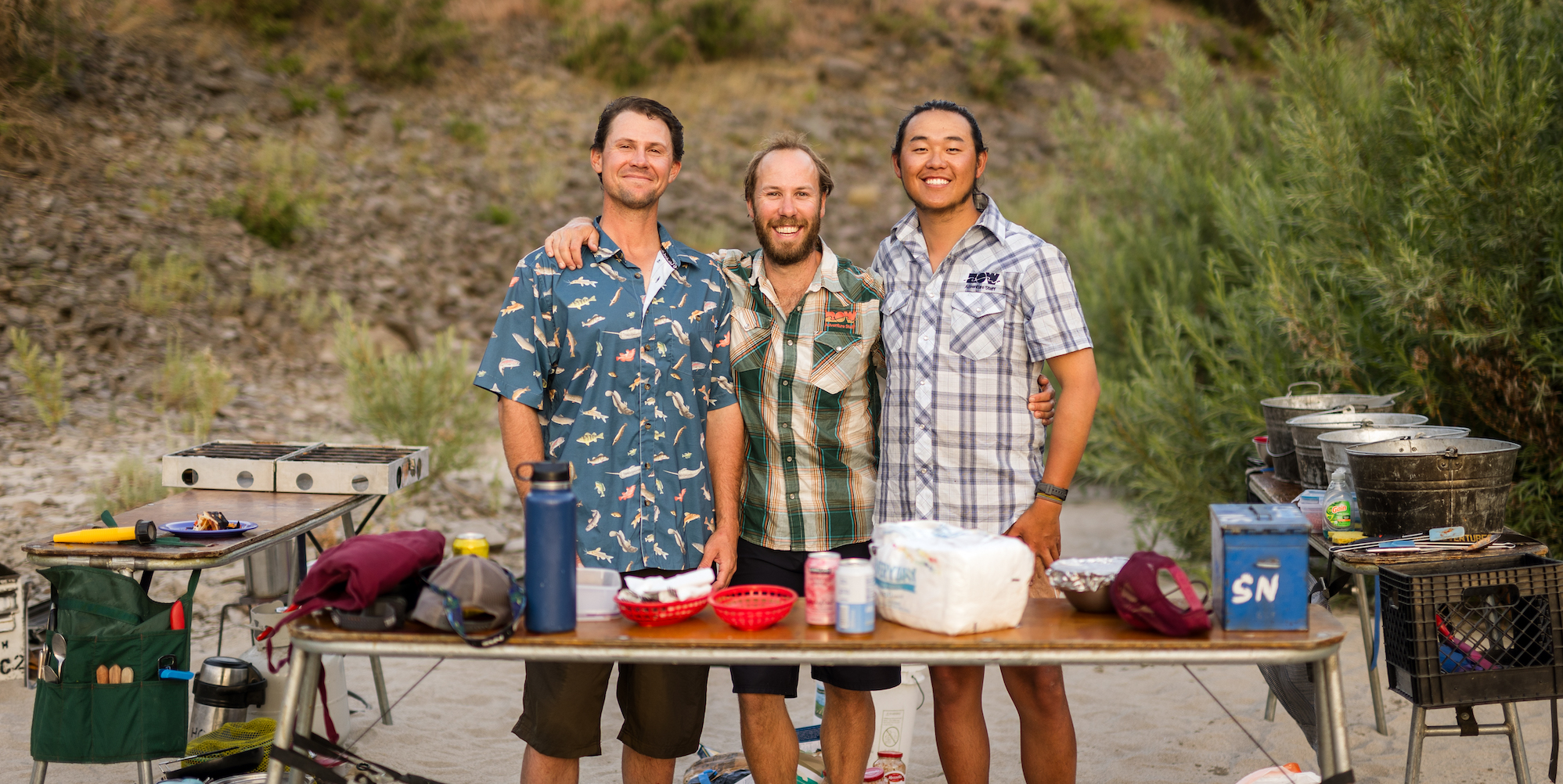 Three male river guides standing behind a camp kitchen table smiling for the camera