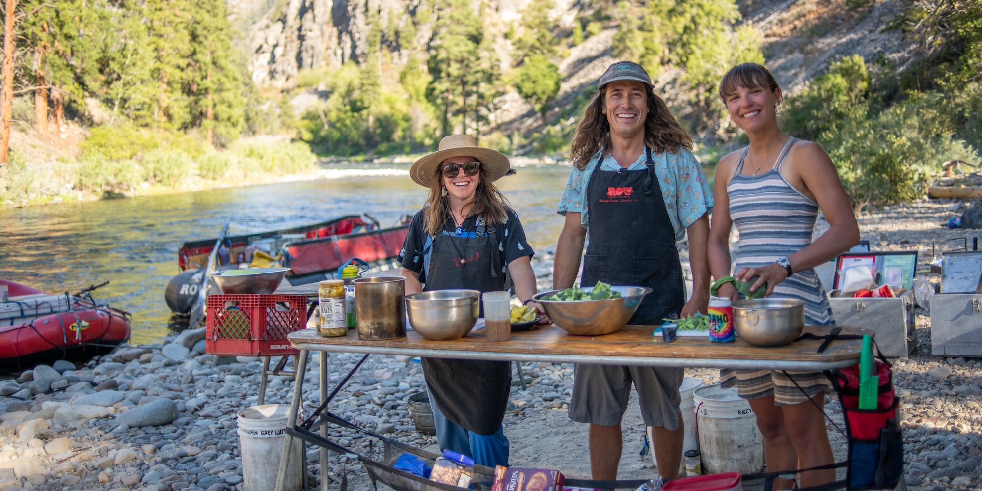 Three river guides smiling while preparing a fresh meal at a riverside kitchen, with ROW rafts and a scenic mountain river in the background