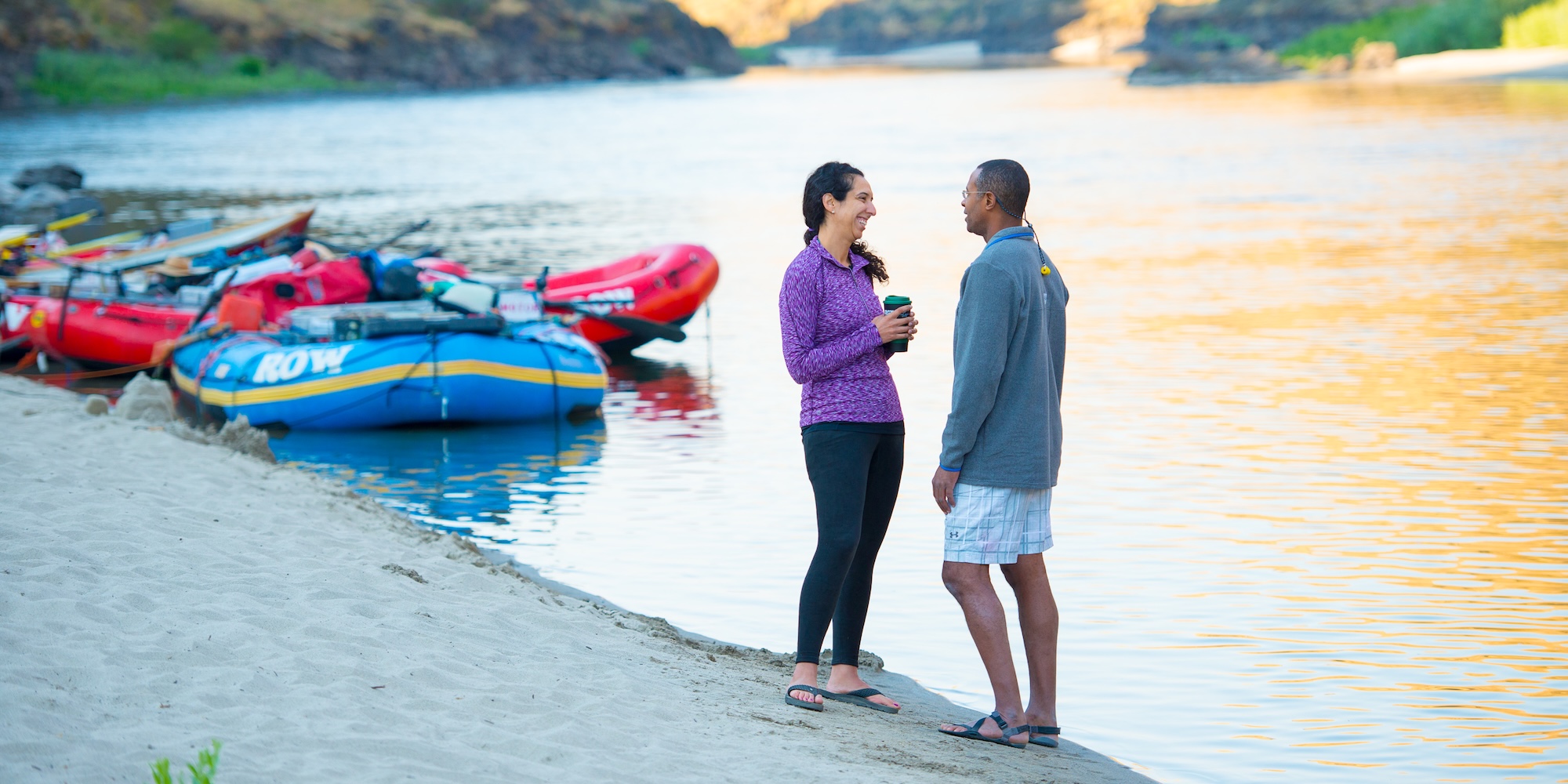 Two people smiling and chatting beside calm river waters at sunrise, with colorful ROW rafts in the background—capturing a quiet morning moment on a river journey.