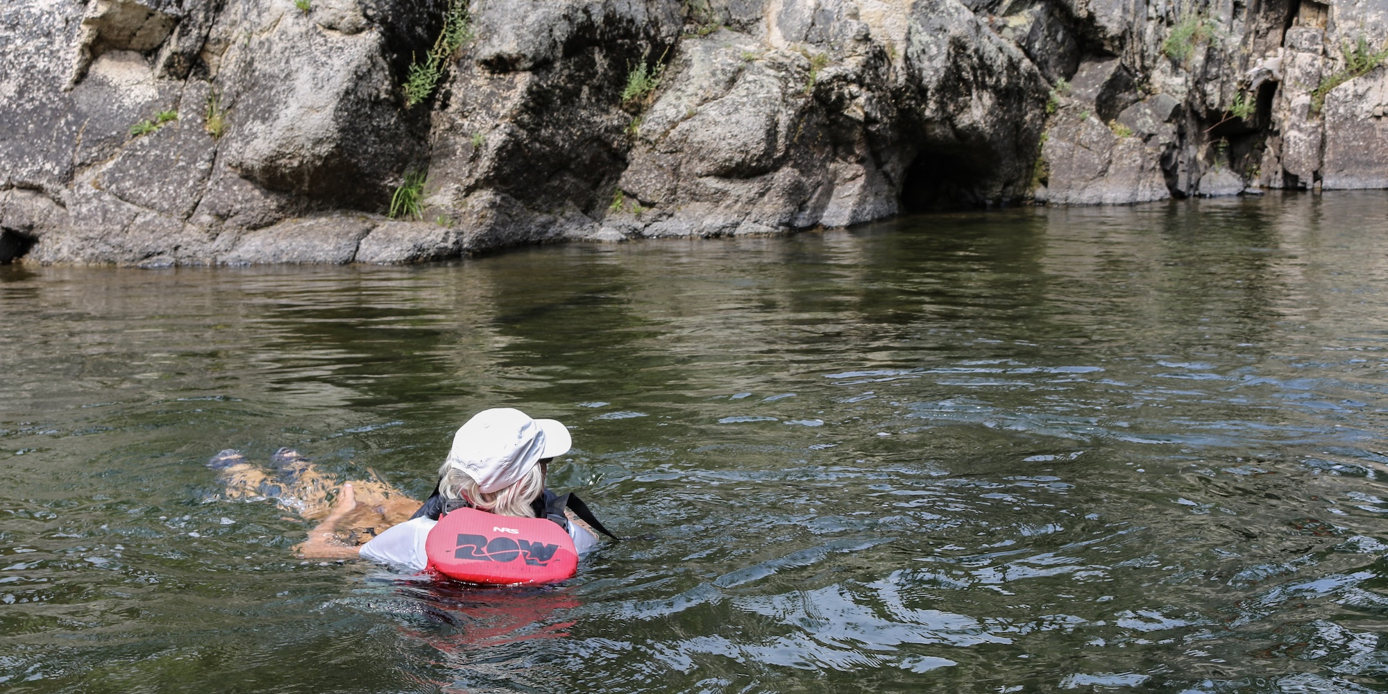 A guest in a ROW life jacket floats peacefully in clear river water, gazing toward rocky cliffs—soaking in the serenity and playfulness of wilderness immersion.