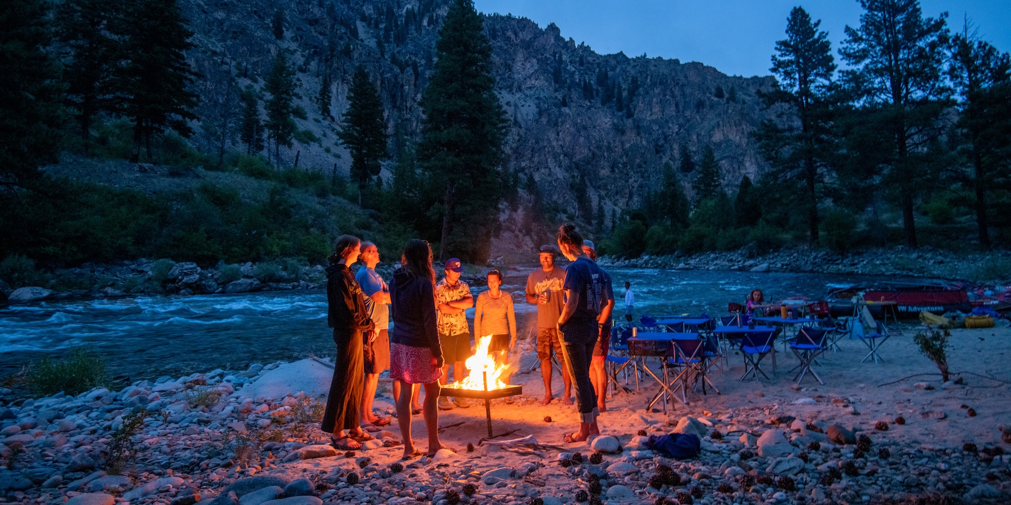 A group of people gathered around a riverside campfire at dusk, sharing stories beneath pine trees and rugged canyon walls—an evening ritual of connection on a multi-day whitewater rafting trip.