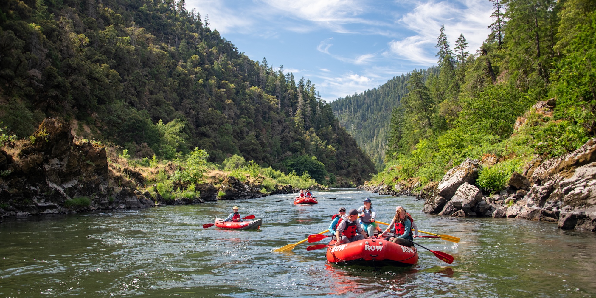 Multiple red rafts float downstream through a lush green canyon, paddlers in sync—immersed in the flow of the river and the joy of shared adventure.